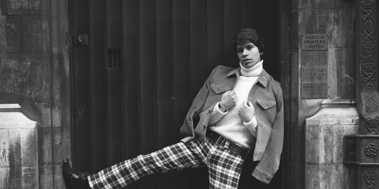 A man is standing in a doorway, wearing chequered trousers, a turtleneck top and a jacket. He has his right leg kicked up in the air. The image is black and white and was taken in the 1960s.