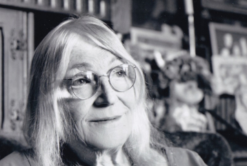 A black and white photo of an older woman, sitting down, with shoulder length hair and glasses. She's looking into the distance.