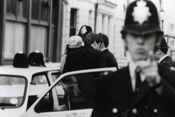 A policeman is taking three people into their police car. In front of the car is a policeman pointing aggressively to the person behind the camera.