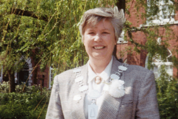 A photograph of Sue King, smiling. She has short hair and is wearing a grey plaid suit.