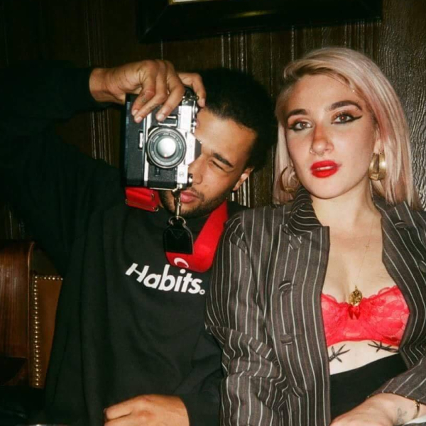 The two people who run London Vagabond. The man on the left is taking a photo, and the woman is sitting next to him, both looking at the camera.