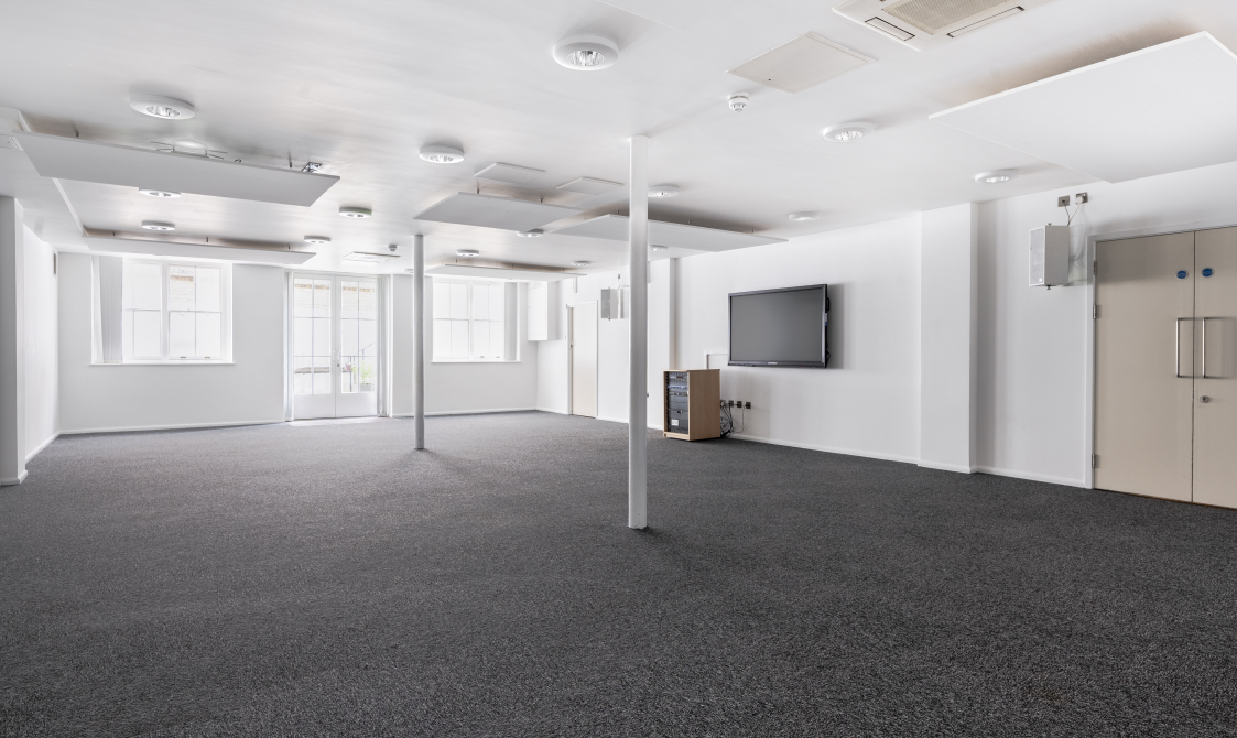 A photograph of Bishopsgate Institute's Courtyard Room in central London. The floor is carpeted and the walls are white, with a screen mounted to the walls.