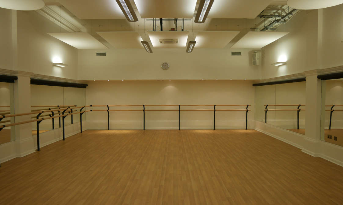 A photograph of the Studio. The floor is wooden and the walls are white, with dance bars around the edge of the room and mirrors on the walls.