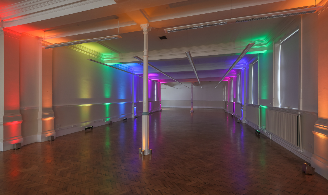 A photograph of the Upper Hall, which has laminate flooring and white walls. Rainbow coloured uplighters are on.