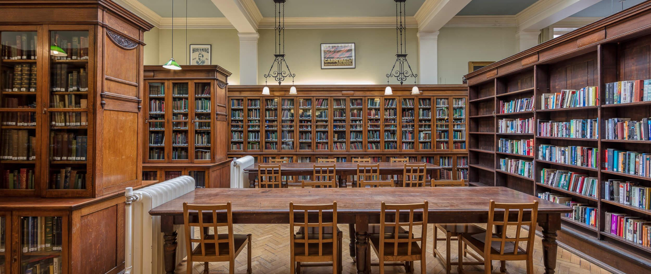A photograph of a London-based Library. There is a wooden table in the middle of the room with rows of bookshelves around it. There is a dome in the ceiling letting light through.