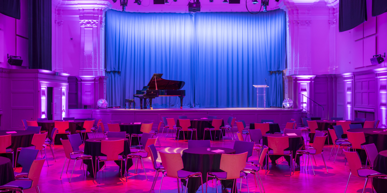 A photograph of the Great Hall, with seating in a cabaret style. There is a stage with a piano on it and blue curtain. There is a purple wash over the room.