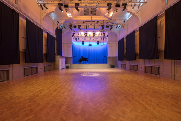 A photograph of the Great Hall. There is a piano on stage with a blue curtain. There is a lighting rig with soft lighting over the stage.