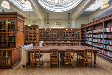 A photograph of a London-based Library. There is a wooden table in the middle of the room with rows of bookshelves around it. There is a dome in the ceiling letting light through.