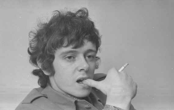 Black and white photo of Scottish singer Donovan. He's biting his thumb whilst smoking a cigarette, looking off in the middle distance.
