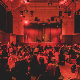 A large group of people sat on the floor of the Great Hall, watching two speakers who are seated on stage