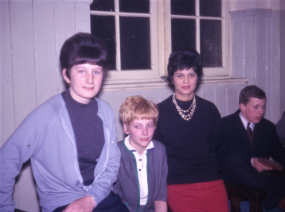 Archive photo with three people posed facing the camera, and a fourth sitting in the background. They are indoors, against a window which shows that it is evening and dark outside.
