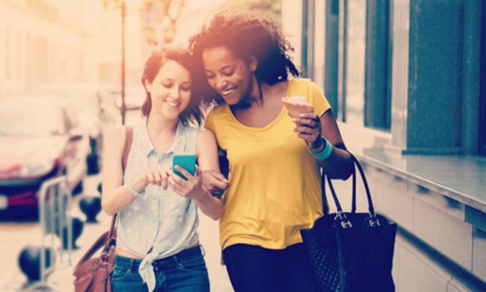 Two girls laughing over a mobile phone, walking down the street. Explore the Lincoln Big Hen Hunt for your hen weekend: