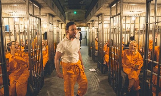 A group of 10 People in orange jumpsuits singing in cells.