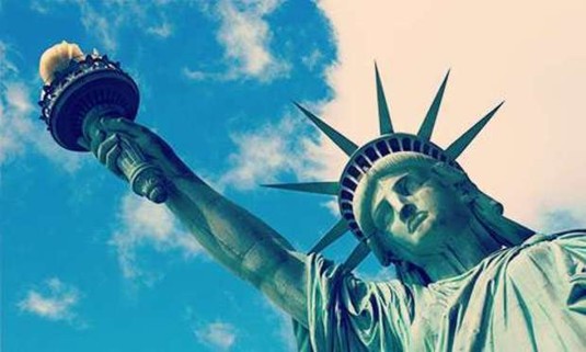 Looking up towards the crown and torch of the Statue of Liberty against a cloudy, blue sky. Discover New York Hen Party ideas below: