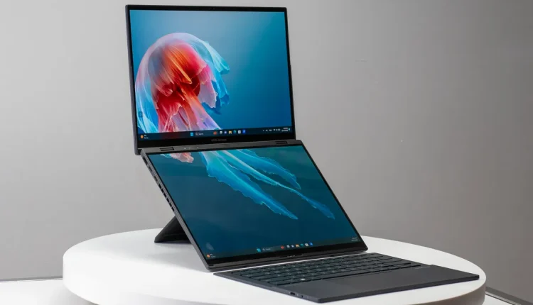 Dual-screen Asus laptop placed on a round white table, with the main display tilted upward and a secondary screen positioned above the keyboard
