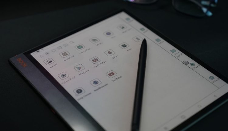 An overhead, angled shot of a Boox Note Air3 C e-reader tablet with its screen displaying a grid of application icons, some of which are labeled in Arabic (e.g., "متجر التطبيقات", "YouTube", "Play Store"). A black stylus pen is lying diagonally across the screen. The device is on a dark surface, with part of a pair of glasses visible in the background.