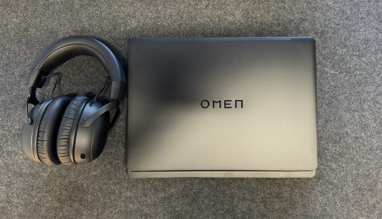 A top-down view of a closed dark grey or black HP Omen laptop with the "OMEN" logo centered on its lid, resting on a dark grey textured surface. To its left, a pair of black HyperX Cloud III over-ear gaming headphones are placed.