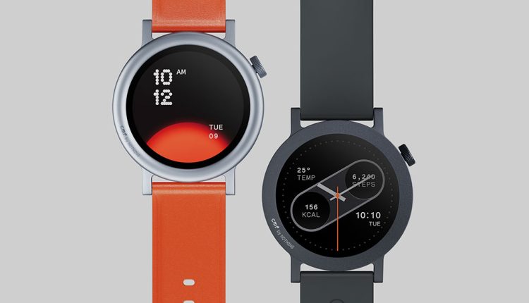 A close-up view of two CMF Watch Pro 2 smartwatches side-by-side on a light grey background. The watch on the left has a silver bezel and a bright orange band, with its black screen displaying the time "10 AM 12" and "TUE 09" against an orange gradient. The watch on the right has a dark grey bezel and a matching dark grey band, with its black screen showing temperature, steps, calories, and the time "10:10 TUE".
