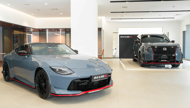 A light blue Nissan Z NISMO sports car is parked in the foreground of a bright car showroom. In the background, a dark gray Nissan Patrol NISMO SUV is displayed on a platform in front of a wall with the NISMO logo.