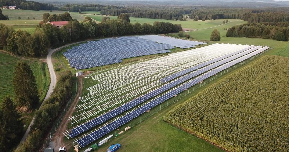 Aerial view of a solar farm under construction, with panels on some racks and others still empty, beside farmland and forest
