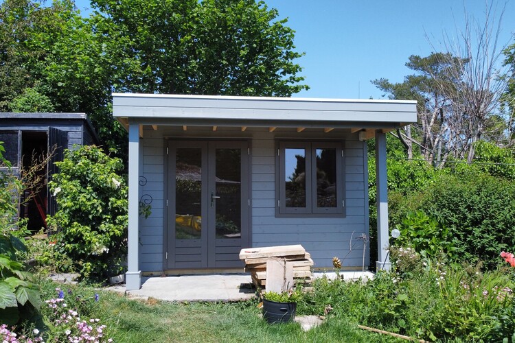 A small, light blue garden office with double doors stands amidst lush greenery. A stack of wooden planks and a black pot rests on the grass in front, surrounded by vibrant plants and flowers. The clear blue sky overhead completes this picturesque scene.