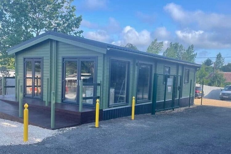 A charming green Lugarde twin cabin building with large windows rests on a gravel lot, boasting a dark base and a wooden deck in front. Its surrounded by yellow bollards, framed by trees and a blue sky. This bespoke structure offers unique accommodation options, with cars visible to the right.