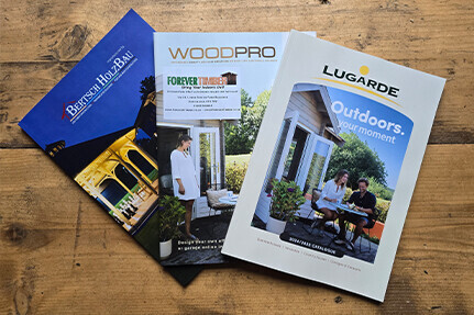 Three brochures are laid out on a wooden surface. The brochures, titled Bertsch Holzbau, Woodpro, and Lugarde, feature images of outdoor structures like log cabins and garden offices set against picturesque scenes.