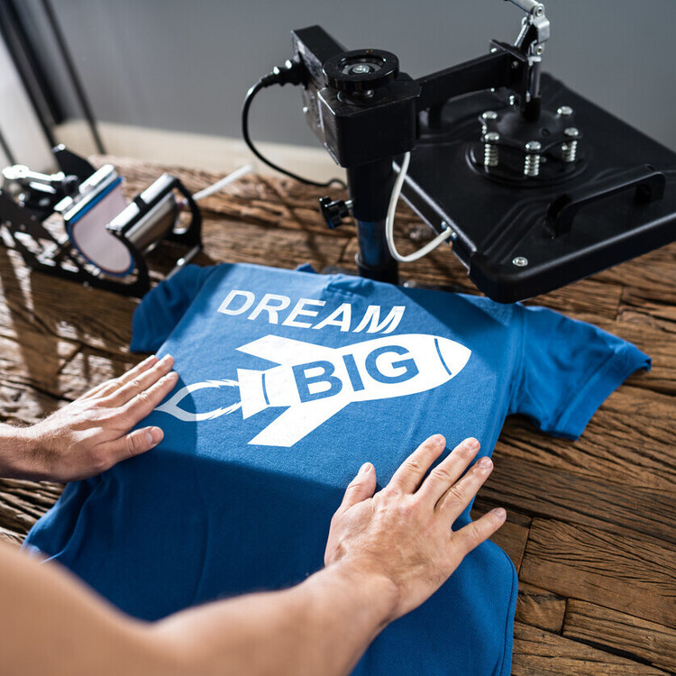 Heat press applying “Dream Big” rocket design onto blue t-shirt, example of vinyl heat transfer (HTV) eco apparel printing.