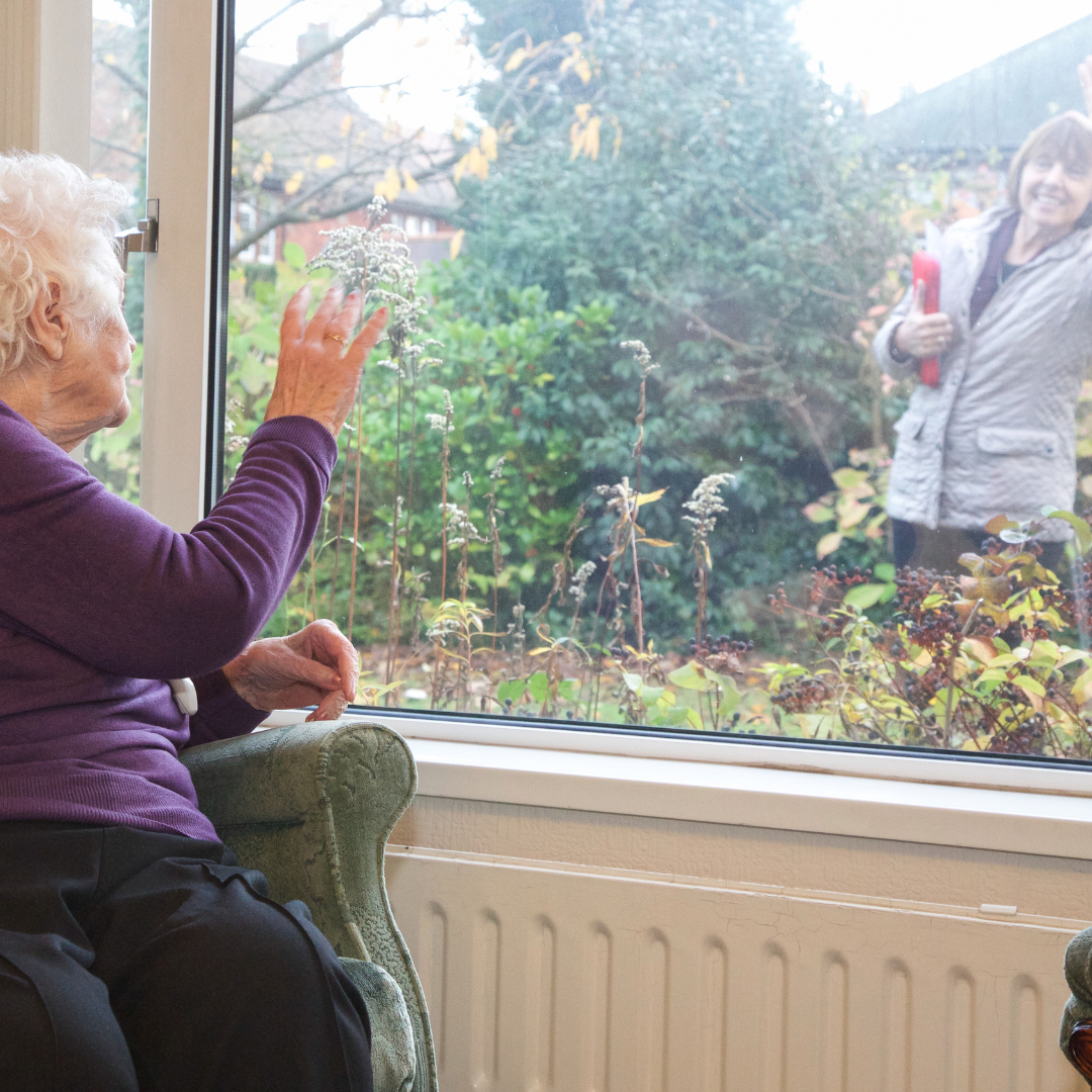 Elderly woman smiling and waving from her window, looking directly at her carer outside on the garden path — a warm, genuine goodbye after a Your Care visit