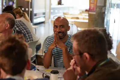 A smiling man sat among colleagues at a busy workshop table.