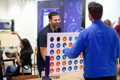 Two people laughing and playing a large tabletop Connect Four game at a conference, with other attendees socialising in the background.