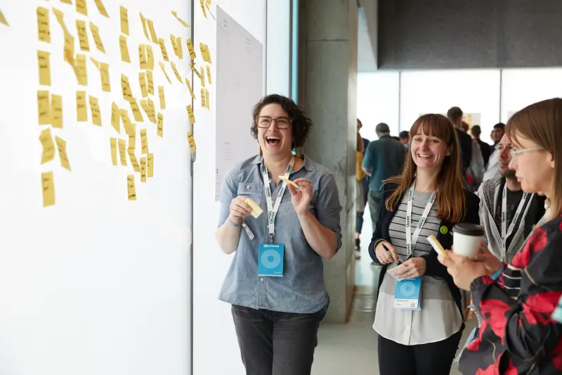 3 women stood laughing in front of a bright window adorned with sticky notes.