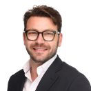 Smiling man with glasses in a black suit and white shirt, set against a plain white background. He exudes a friendly and approachable vibe.