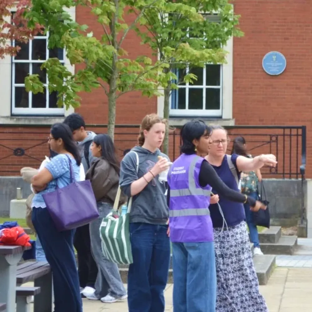 Visitors gather outside a red-brick University of Manchester building on an open day. A student guide in a purple hi-vis vest points the way for a group, while a purple "Campus tours" flag stands nearby.