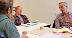 Three people sitting around a table in a workshop or meeting, discussing a card-based activity with notebooks and coffee cups.