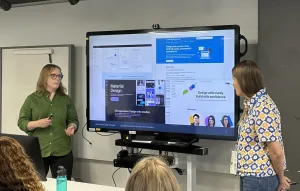 Two presenters stand on either side of a large screen displaying examples of design system interfaces. The presenter on the left is speaking and gesturing with a clicker, while the presenter on the right listens. Several audience members are seated in front of them.