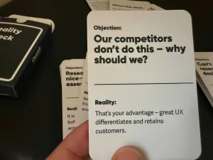 Close-up of a hand holding a card that reads, “Objection: Our competitors don’t do this — why should we? Reality: That’s your advantage — great UX differentiates and retains customers,” with other cards and a card deck visible in the background.