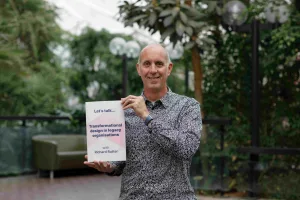 A person standing outdoors holding a sign that reads “Let’s talk… Transformational design in legacy organisations,” with greenery and seating visible in the background.
