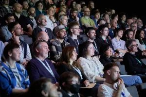 Audience seated in a conference auditorium, attentively watching a presentation, with many wearing lanyards and focused expressions.
