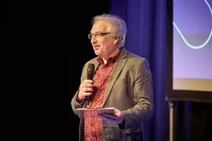 A man stands on stage holding a microphone and a small tablet or notes, speaking to an audience, with a presentation screen behind him.