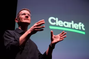 A dramatic shot looking up at Chris on stage. On screen behind him is the Clearleft logo.