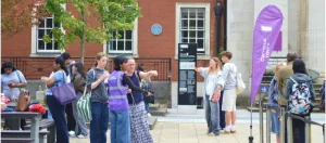 Visitors gather outside a red-brick University of Manchester building on an open day. A student guide in a purple hi-vis vest points the way for a group, while a purple "Campus tours" flag stands nearby.