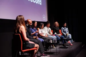 Day 1 speakers seated in a row on a stage, holding microphones and talking under soft stage lighting, with a projected screen behind them.