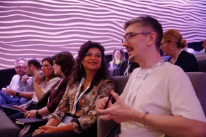 Attendees seated in a row inside the auditorium, chatting and smiling under soft purple lighting with a wavy textured wall behind them.