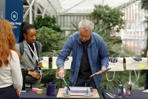 A person demonstrates screen-printing at a table while attendees watch, set against lush indoor greenery and hanging printed tote bags.