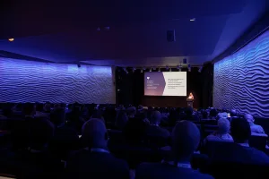 Audience seated in a dark auditorium watching a presenter speak on stage, with blue lighting and textured wavy walls framing the room.
