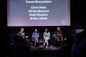 A four-person panel seated on stage in front of a screen listing the speakers’ names, with a moderator holding notes and the audience watching.
