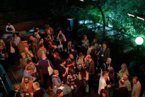 A large group of attendees gathered in a lush indoor garden space, mingling and chatting during a networking reception.