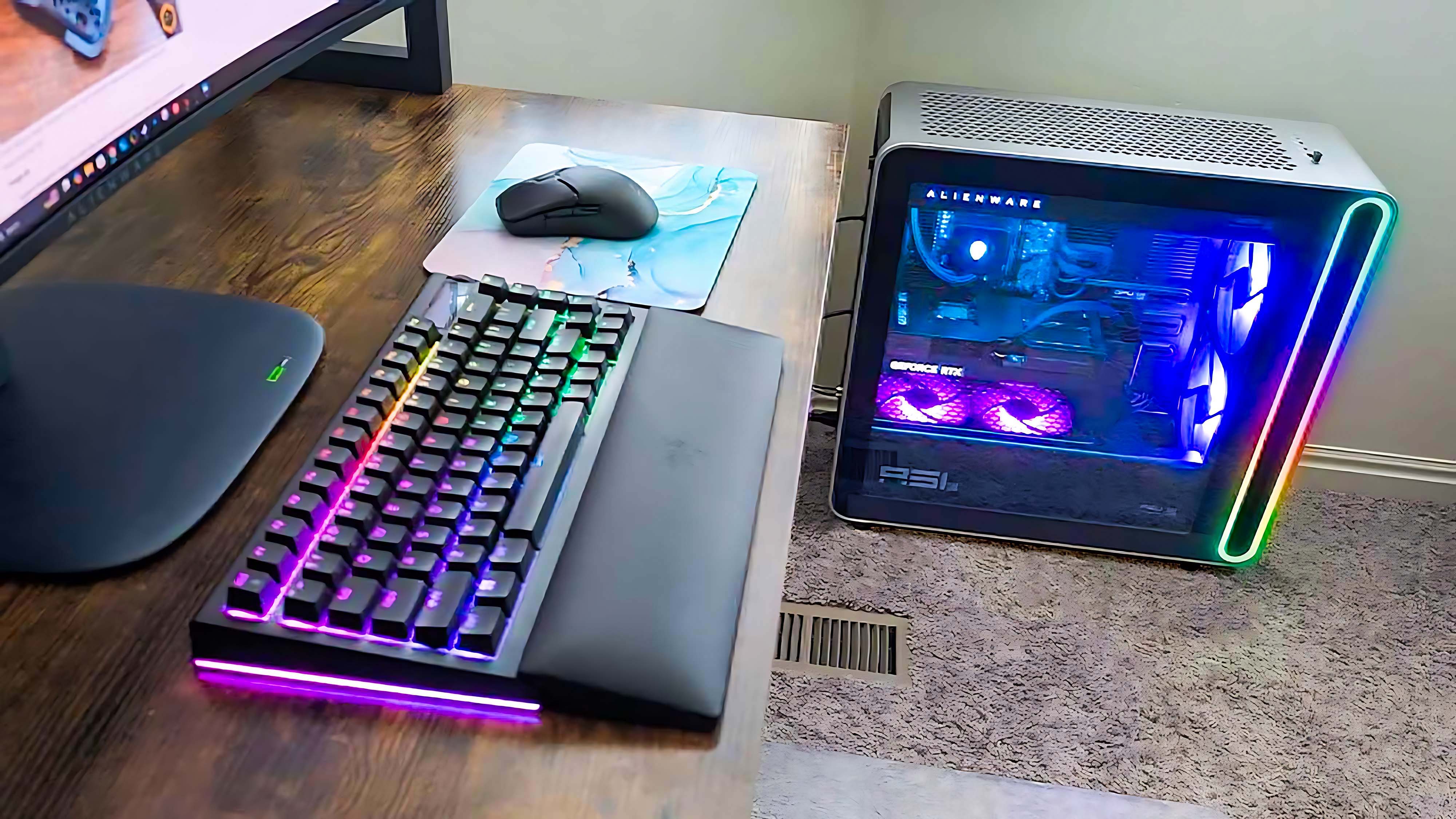 Вид сбоку на стол, на котором видны и RGB-клавиатура, и игровой компьютер Alienware Area-51 Gaming Desktop (2025).