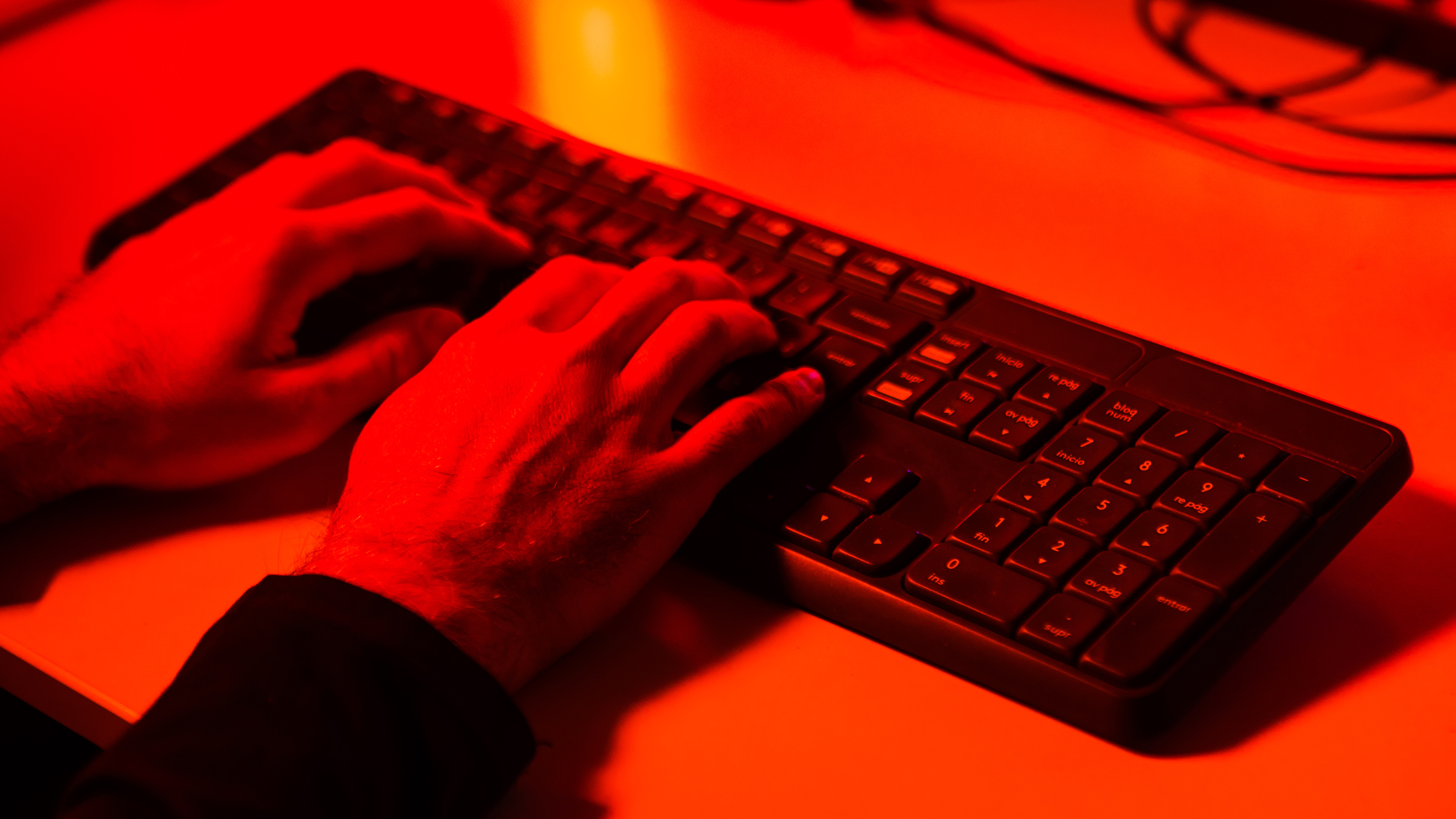 Руки человека, набирающие текст на клавиатуре компьютера, освещенные красным светом. Концепция кибербезопасности, кражи, хакера, личности и преступления.
